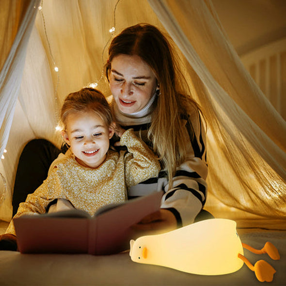 Veilleuse canard allumée illuminant une maman lisant un livre avec son enfant sous un ciel de lit, créant une ambiance magique. L'image dépeint la douceur et l'atmosphère enchantée que la veilleuse apporte lors des moments de lecture avant le coucher.