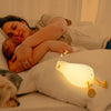 Veilleuse canard allumée placée sur un lit, à côté d'une maman tenant fermement son bébé dans ses bras, tous deux dormant paisiblement. L'image illustre le confort et la sécurité que la veilleuse apporte pendant le sommeil.