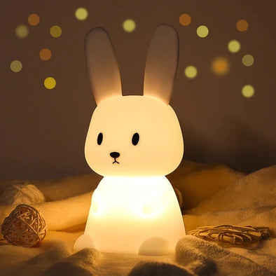 Veilleuse en forme de lapin sur un lit illuminée d'une couleur chaude dans une ambiance féerique.